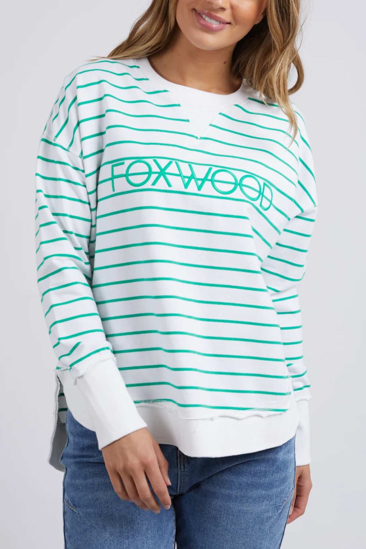 A woman wears the Foxwood Simplified Stripe Crew, a relaxed white 100% cotton sweatshirt with green stripes and "FOXWOOD" on the front, paired with blue jeans.