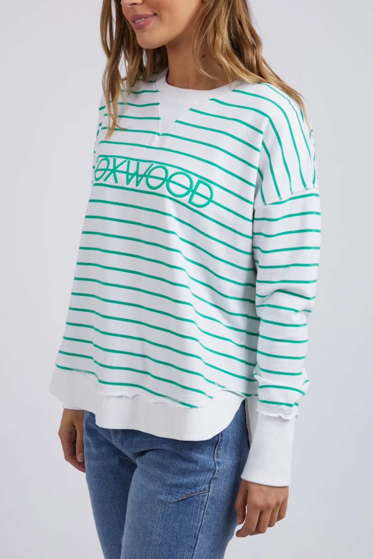 A woman models the Foxwood Simplified Stripe Crew, a white cotton sweater with green stripes and "OAKWOOD" text, paired with blue jeans. She stands before a light backdrop, highlighting the sweater’s relaxed fit.
