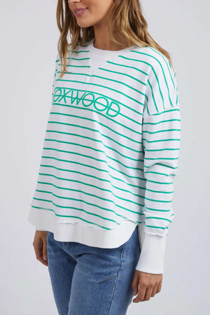 A woman models the Foxwood Simplified Stripe Crew, a white cotton sweater with green stripes and "OAKWOOD" text, paired with blue jeans. She stands before a light backdrop, highlighting the sweater’s relaxed fit.