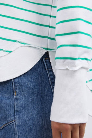 A close-up of a person wearing the Foxwood Simplified Stripe Crew, crafted from 100% cotton and featuring green stripes, layered over a white shirt. The ensemble is paired with blue jeans, showcasing the sweater's relaxed fit and textures. The image focuses on the side of the sweater and hand to emphasize its vibrant colors.