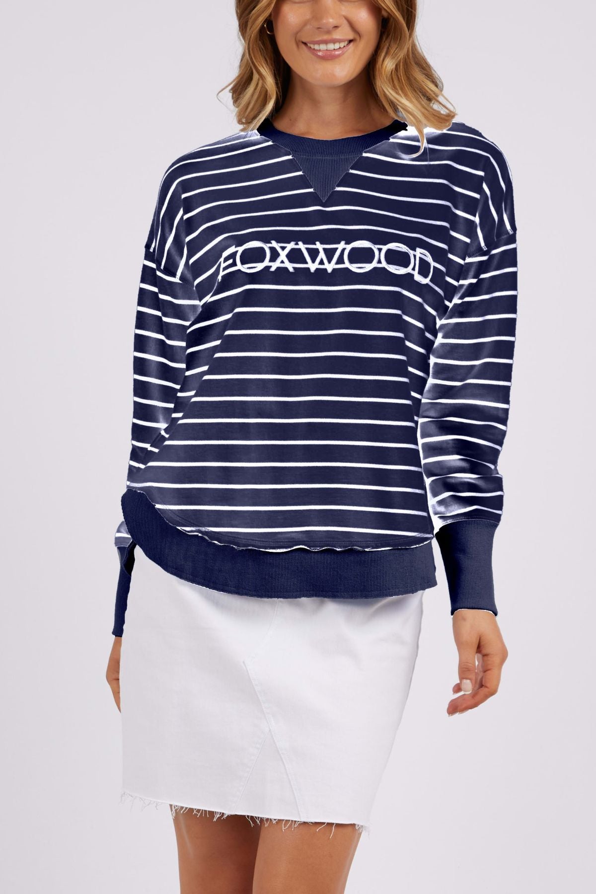 A person is sporting a navy blue and white striped "Foxwood Classic Stripe Simplified Crew" sweater, crafted from 100% cotton, with a relaxed fit and "FOXWOOD" emblazoned on the front. They have paired it with a white skirt featuring a frayed hem against a plain light gray backdrop.