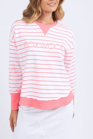 A person is wearing the Foxwood Simplified Stripe Crew, a stylish sweatshirt with white and pink stripes and "FOXWOOD" emblazoned on the front. This layering option from Foxwood Clothing also features distinctive pink cuffs and a neckline. Paired with a white skirt, this ensemble stands out against a plain white backdrop.