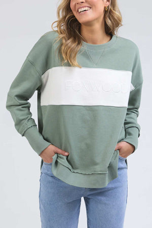 A person in a green Foxwood Simplified Tonal Crew sweatshirt with a white stripe and "FOXWOOD" embossed on it smiles, their long hair cascading down. Their hands rest in the pockets of light blue jeans, while the cotton fleece fabric shows subtle raw edge detailing.
