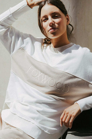 A person with long hair, appearing content and looking towards the camera with a slight smile, is sitting and leaning against a wall while wearing a relaxed, oversized Foxwood Simplified Tonal Crew featuring front chest embroidery.