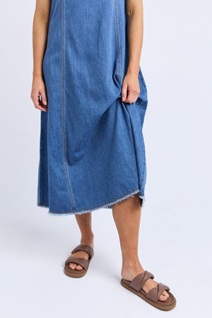 A person wears the Foxwood Solene Denim Dress—a sleeveless blue denim dress with raw hem and A-line silhouette—holding one side. They pair it with brown slide sandals against a plain white background.
