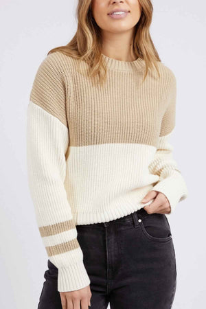 A person with long hair is wearing the Foxwood Sophie Knit Crew, a relaxed-fit beige and white color-block sweater featuring striped sleeves, paired with dark jeans. They are standing against a plain background with one hand partially in their jeans pocket.