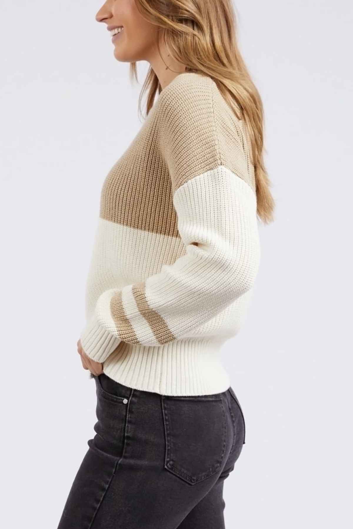 A woman with long, wavy hair stands sideways, smiling. She is wearing the Foxwood Sophie Knit Crew with beige and white horizontal sections and dark grey jeans. The background is plain and light-colored.
