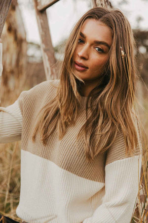 A woman with long, light brown hair stands outdoors near a wooden ladder, wearing the Foxwood Sophie Knit Crew in a relaxed-fit beige and white design. She looks slightly to the right with a calm expression, and the background shows blurred natural scenery. The sweater features subtle color blocking for added style.
