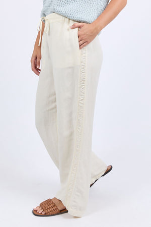 A woman wears the Foxwood South Pantβlight beige, wide-leg linen pants with a drawstring waist and ruffled trimβpaired with brown woven sandals and a light blue knit top for effortless style and comfort.