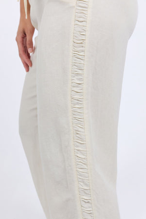 A close-up of a person wearing Foxwood South Pant in light beige, featuring an elastic waist and ruched side seam. The person's hand rests by their side against a plain, light background.