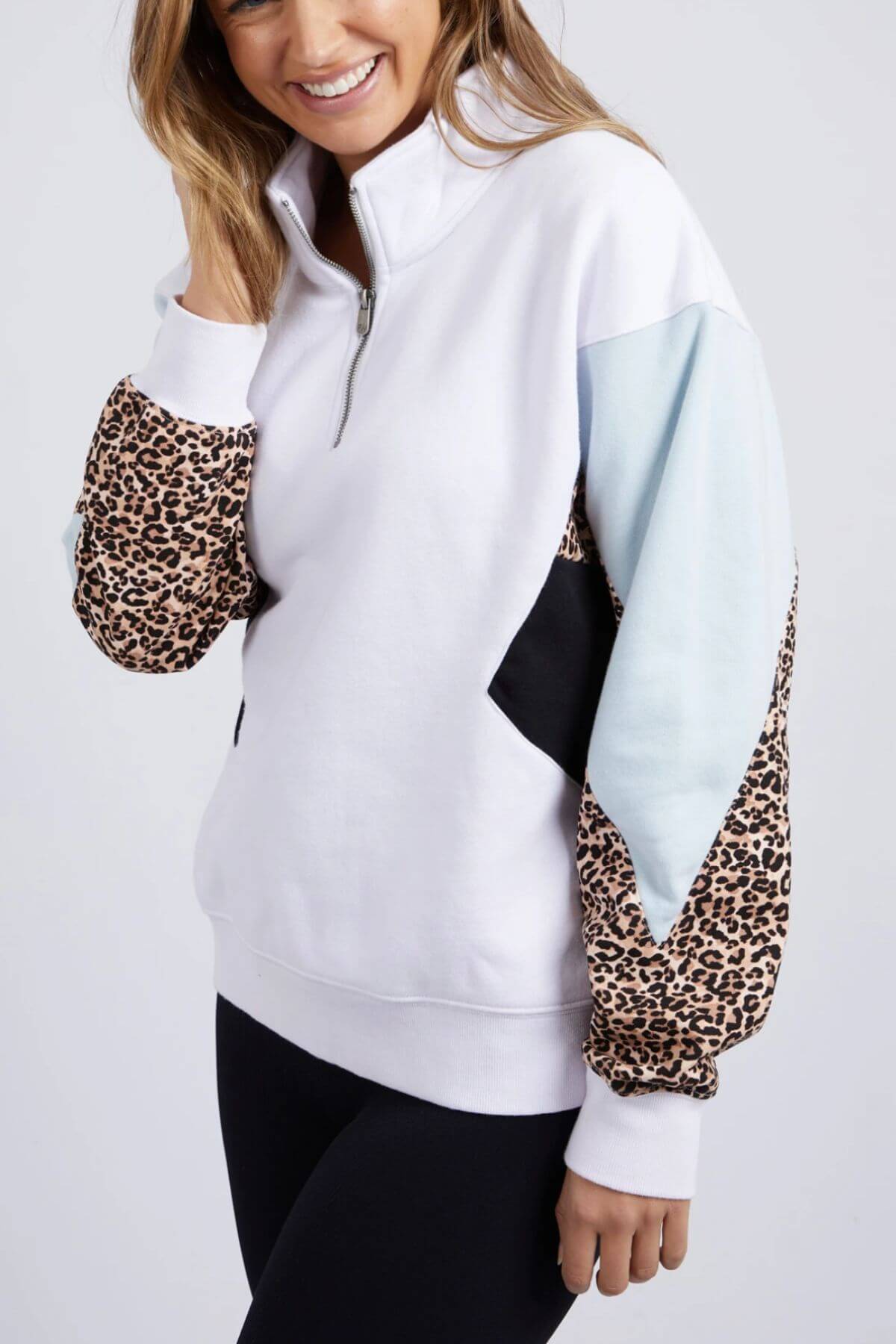 A woman smiles wearing the Foxwood Splice Zip Crew, a relaxed-fit cotton pullover with black and light blue color-blocked sleeves and leopard print accents. She has long hair and pairs it with black leggings.