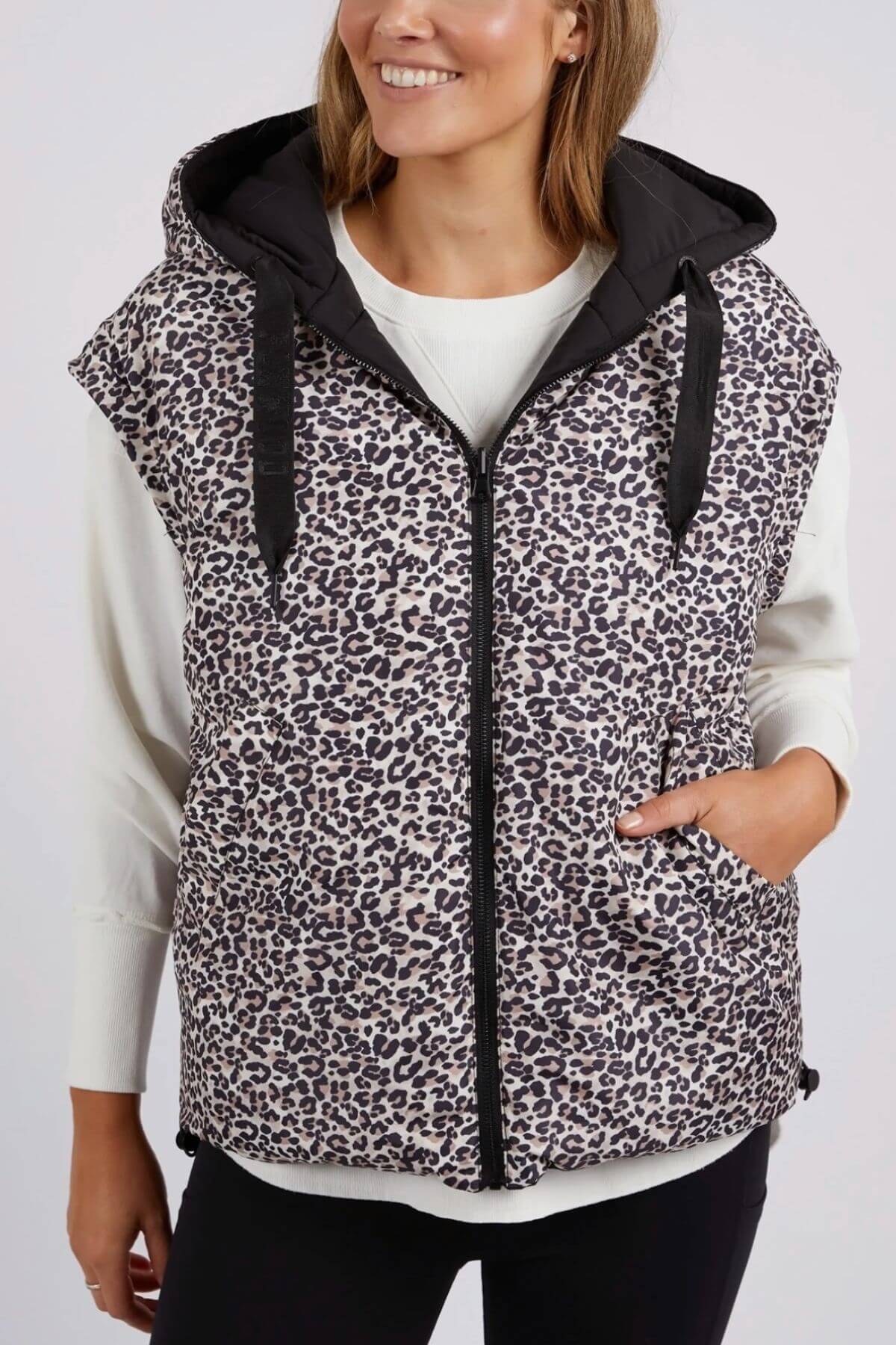 Wearing a sleeveless Foxwood Sports Leopard Vest, a person stands against a plain background. The reversible puffer vest features adjustable toggles and is worn over a white long-sleeve shirt. With one hand in the vest pocket, they smile warmly at the camera.