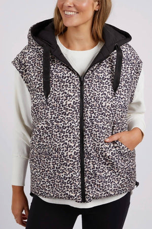 Wearing a sleeveless Foxwood Sports Leopard Vest, a person stands against a plain background. The reversible puffer vest features adjustable toggles and is worn over a white long-sleeve shirt. With one hand in the vest pocket, they smile warmly at the camera.