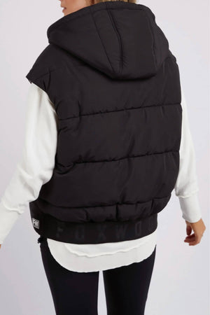 A person in a Foxwood Sports Leopard Vest, black and reversible with a hood and adjustable toggles, over a white long-sleeve shirt stands back to the camera. Only part of the logo is visible on the vest's bottom edge.