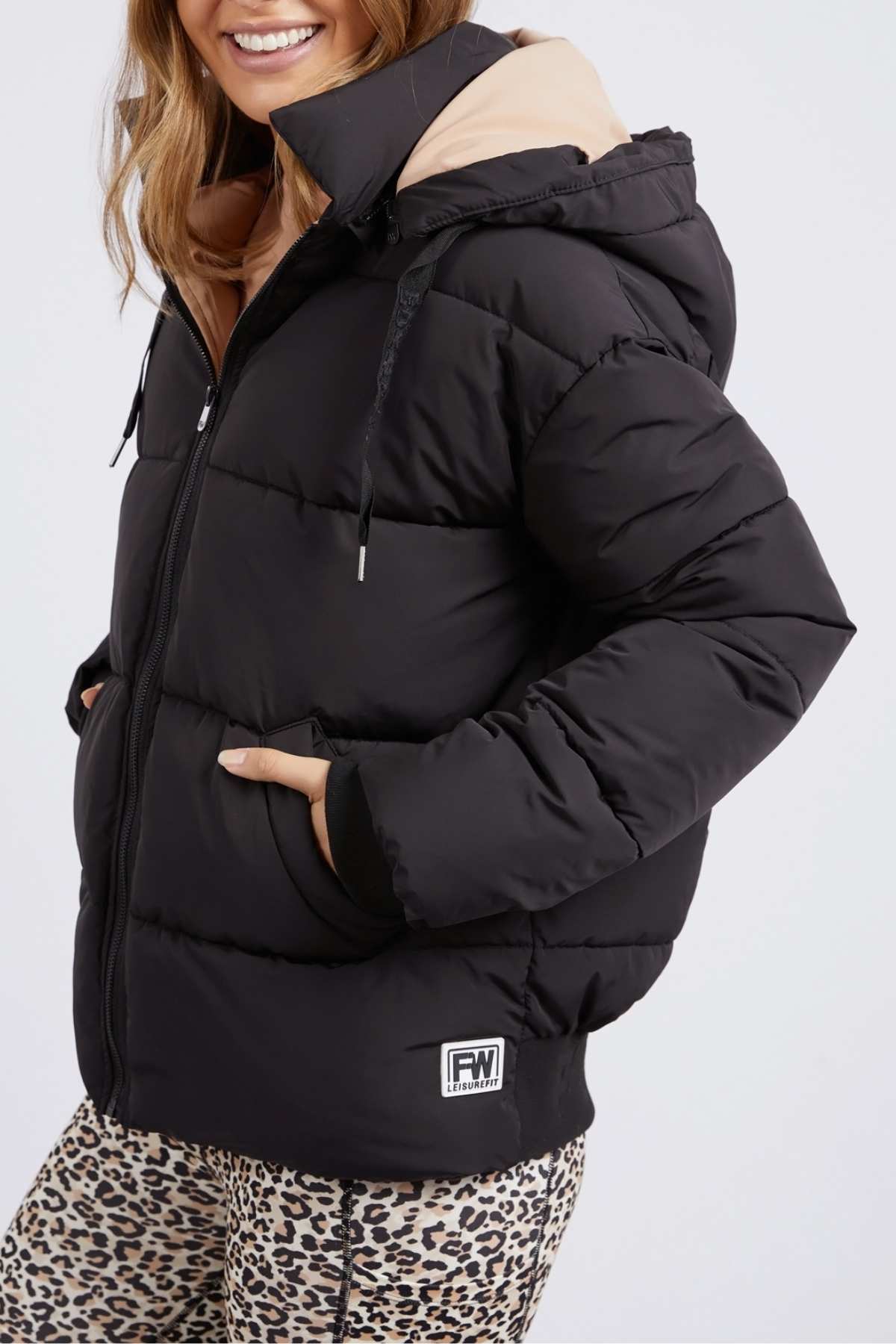 A person is wearing a black Foxwood Sporty Puffa jacket with an oversized hood, paired with leopard print leggings. The versatile jacket has a small logo patch at the hem. They have their hands in the pockets and are smiling, showing only the lower half of their face.