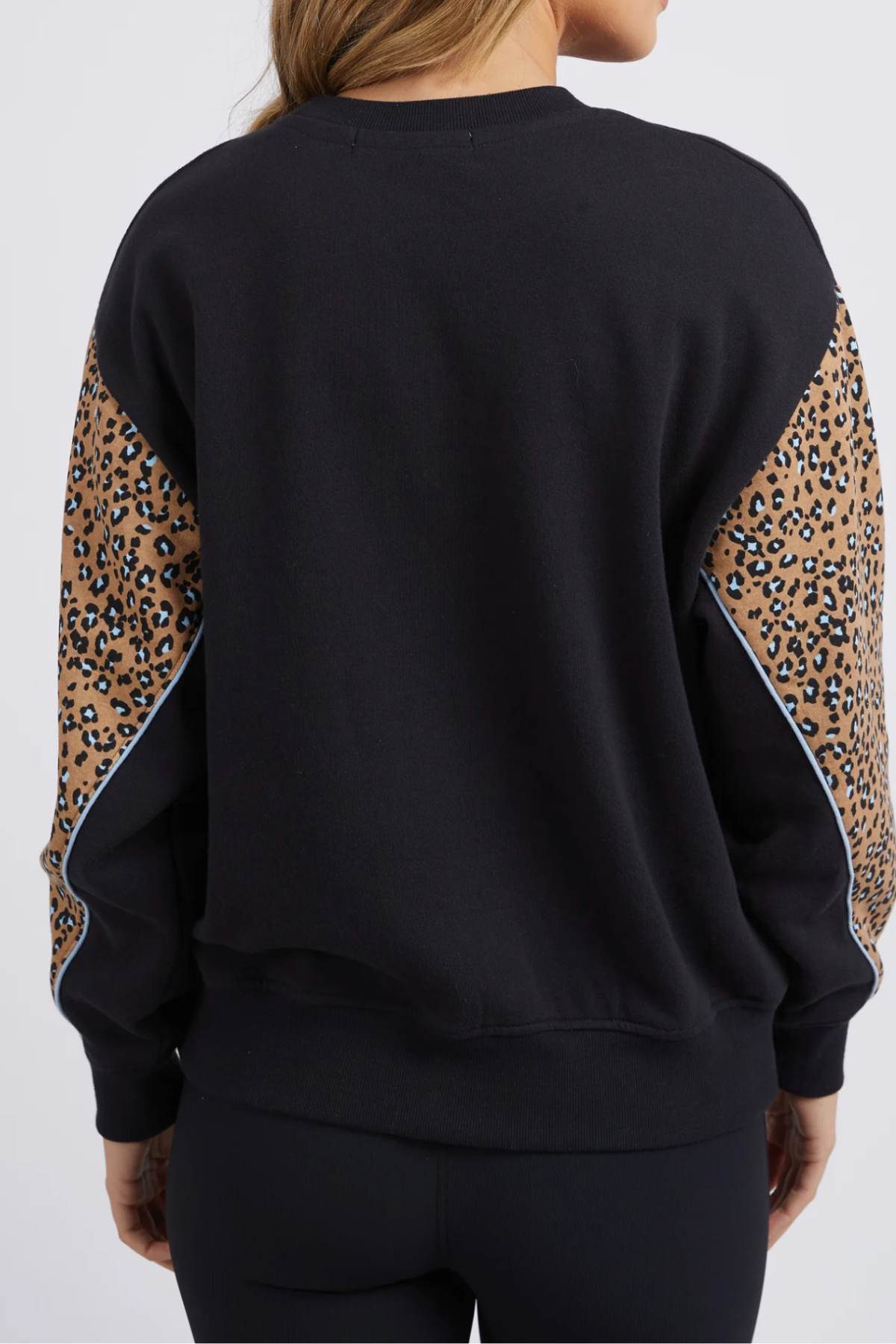 A person with long, light brown hair is shown from the back, wearing the Foxwood The Panel Crew sweatshirt featuring brown and black leopard print panels on the sleeves. They are also dressed in black leggings against a plain white background.
