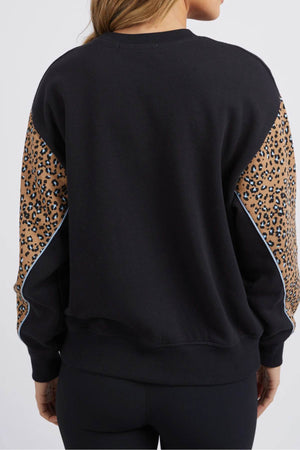 A person with long, light brown hair is shown from the back, wearing the Foxwood The Panel Crew sweatshirt featuring brown and black leopard print panels on the sleeves. They are also dressed in black leggings against a plain white background.