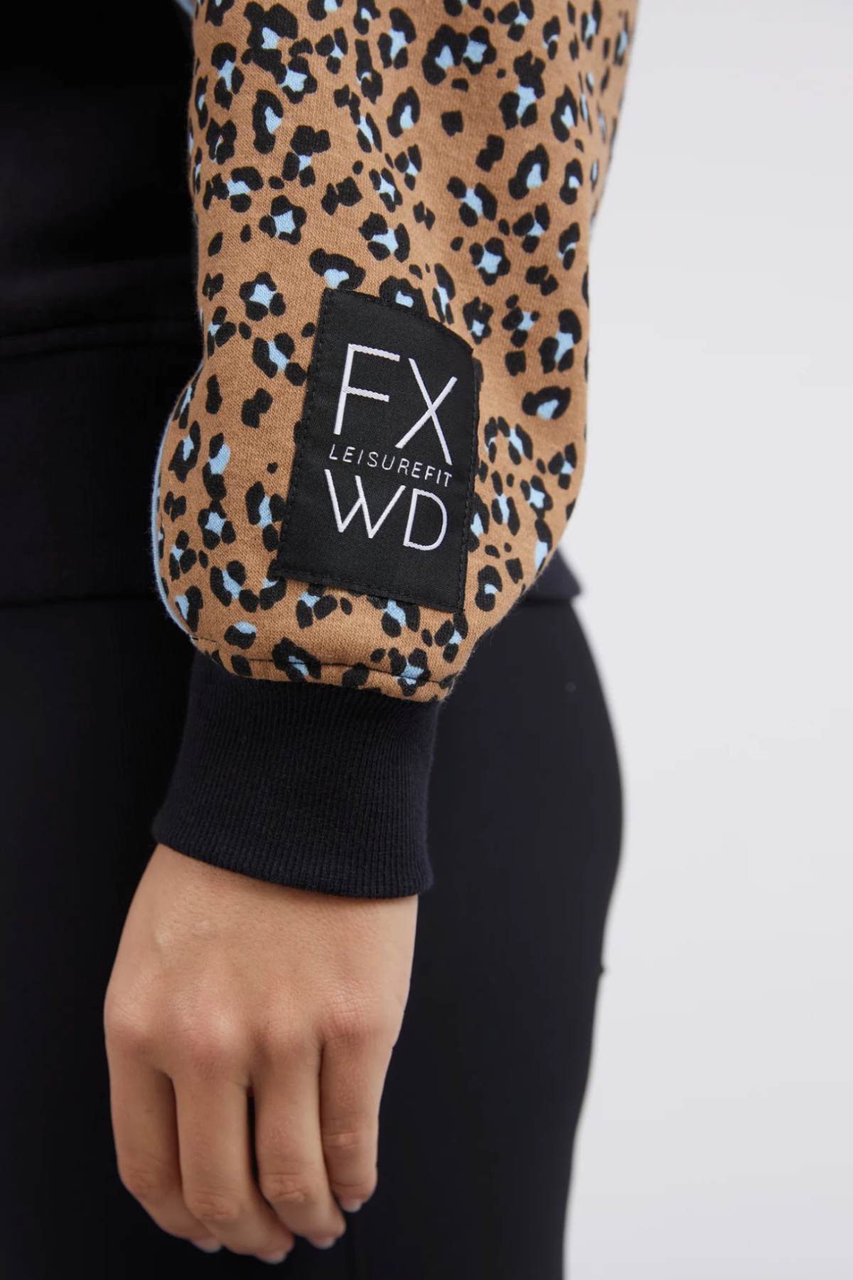 Close-up of a person's arm in a Foxwood The Panel Crew top featuring animal-print panels and a patch labeled "FX WD LEISUREFIT." The sleeve displays a pattern of brown, black, and blue spots. The person is also wearing black pants, with their hand relaxed by their side.