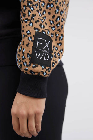 Close-up of a person's arm in a Foxwood The Panel Crew top featuring animal-print panels and a patch labeled "FX WD LEISUREFIT." The sleeve displays a pattern of brown, black, and blue spots. The person is also wearing black pants, with their hand relaxed by their side.