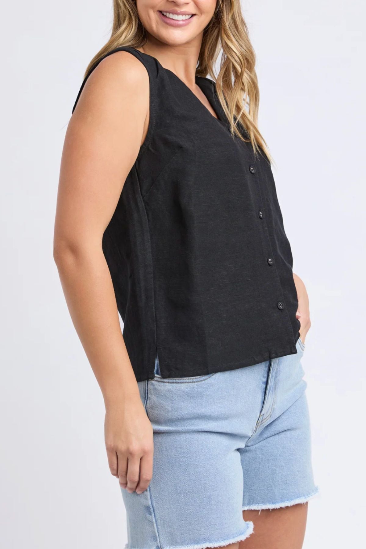 A woman smiles, standing with her hand by her side against a light background, wearing the sleeveless Foxwood Anara Top and light blue denim shorts.