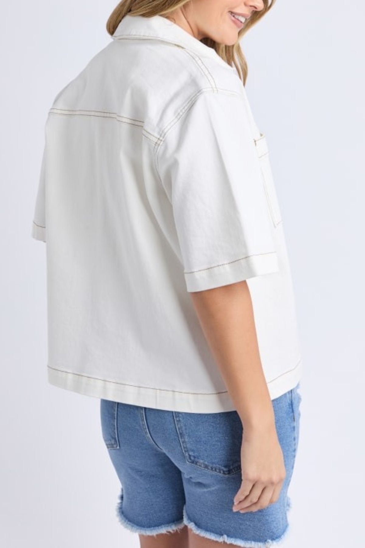 A woman stands sideways, smiling in a Foxwood Valletta Short Sleeve Shirt and blue denim shorts with frayed hems against a plain light backdrop.