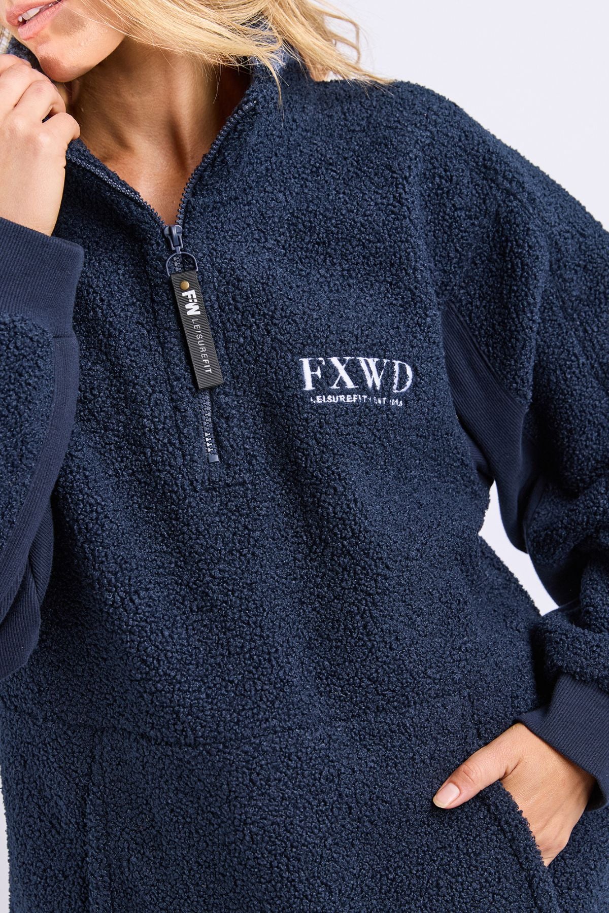 A person wearing the Foxwood Weekend Boucle Crew, a navy blue fleece with "FXWD" and small text embroidered on the chest, cozy half-zip front, and dark ribbed cuffs. Their face is partially out of frame.