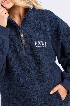 A person wearing the Foxwood Weekend Boucle Crew, a navy blue fleece with "FXWD" and small text embroidered on the chest, cozy half-zip front, and dark ribbed cuffs. Their face is partially out of frame.