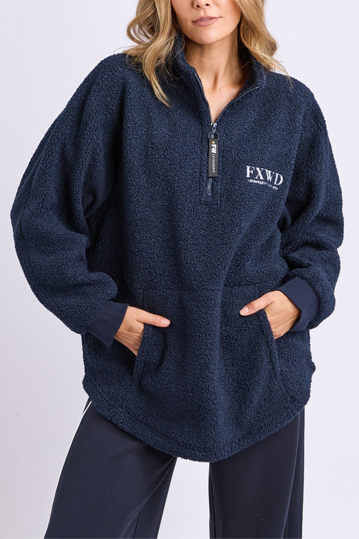A woman wears the Foxwood Weekend Boucle Crew in navy with "FXWD" embroidered on the chest, featuring a cozy 1/4 zip neck and front pocket. She pairs it with matching navy pants for the perfect loungewear look.