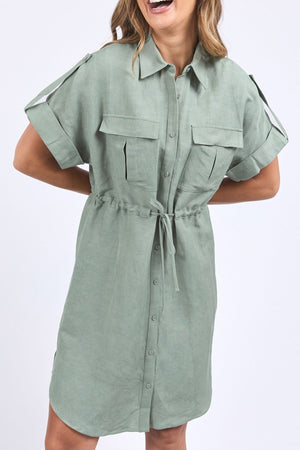 A woman smiles with her hands on her hips, wearing the Foxwood Willow Shirt Dressโa sage green, short-sleeve, button-down dress in a linen-viscose blendโhighlighting its waist tie and chest pockets.