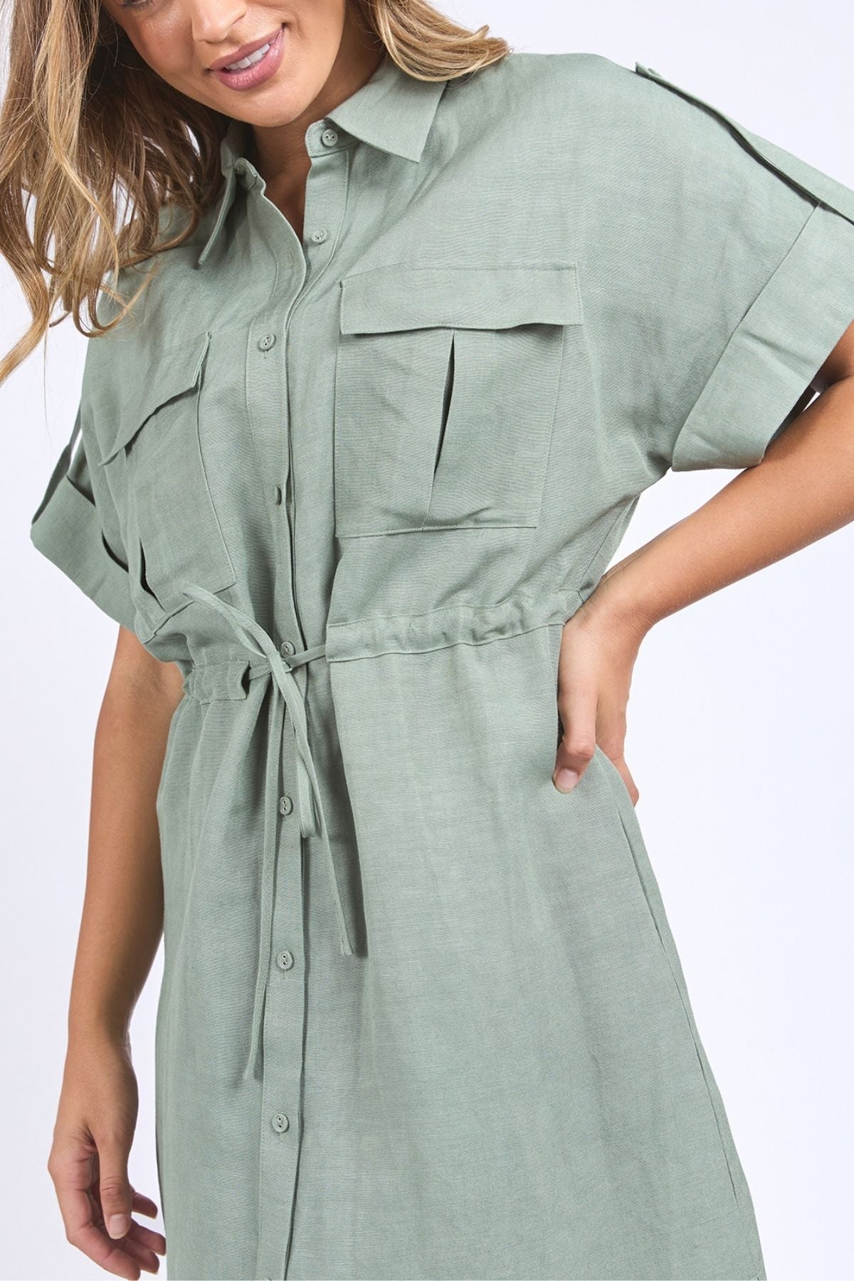 A woman poses with one hand on her hip, wearing the Foxwood Willow Shirt Dressโa sage green, short-sleeved, button-down dress in a linen-viscose blend with a tie waist and two front chest pockets.