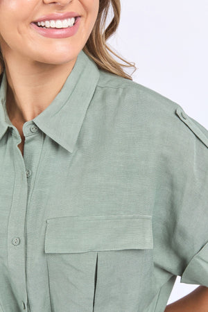 A woman smiles, wearing the Foxwood Willow Shirt Dress in light green linen-viscose, featuring a front pocket and shoulder epaulets, shown from the shoulders up against a light background.
