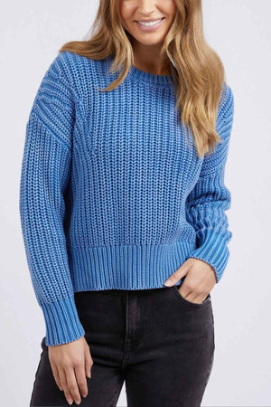 A person with long hair is wearing a cozy blue Foxwood Winnie Knit Crew with black pants, smiling slightly against a plain background. The upper body shot highlights the relaxed-fit jumper's elegant style.