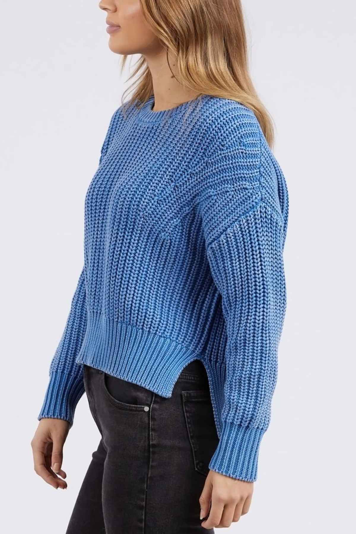The person is in profile against a plain background, donning a textured blue Foxwood Winnie Knit Crew sweater and black jeans. Their long, wavy hair complements the relaxed-fit sweater, which features a side slit at the hem.