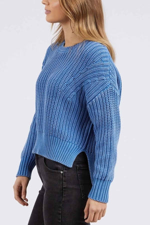 The person is in profile against a plain background, donning a textured blue Foxwood Winnie Knit Crew sweater and black jeans. Their long, wavy hair complements the relaxed-fit sweater, which features a side slit at the hem.