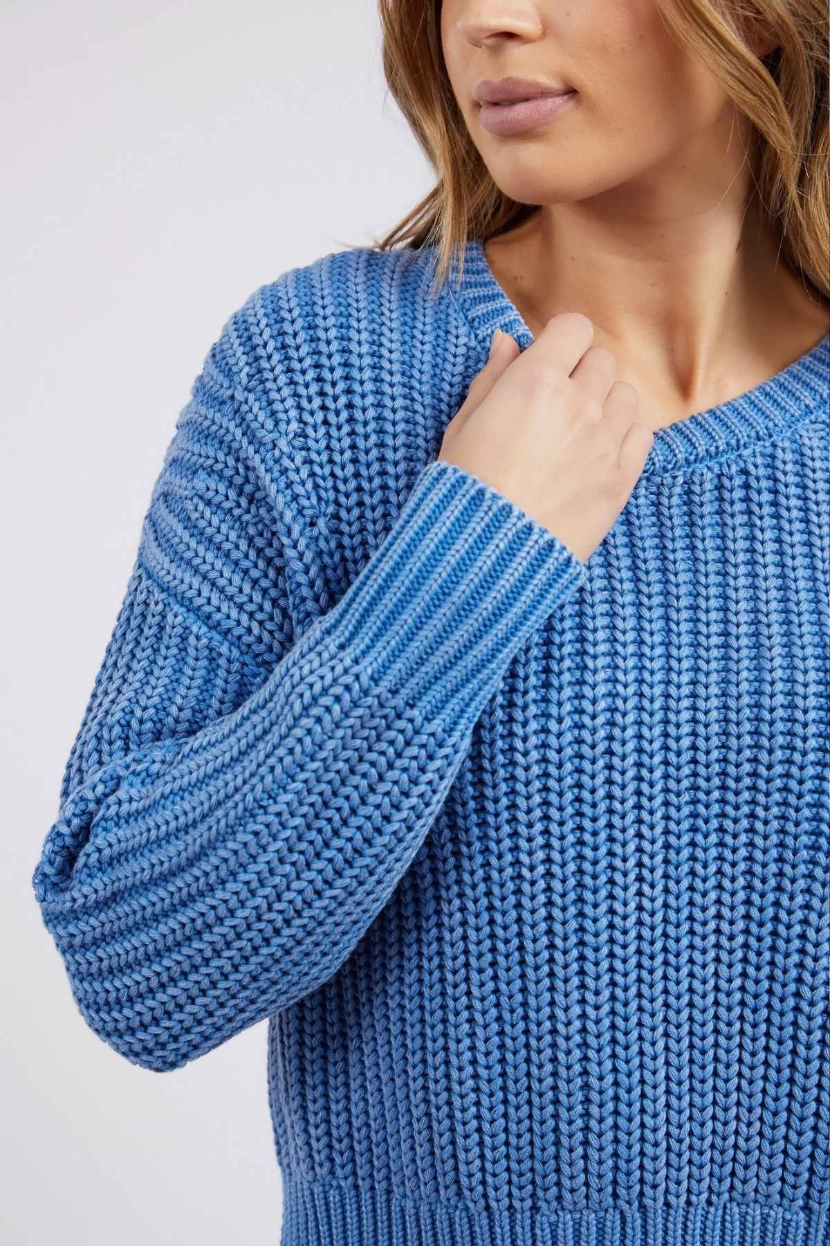In her Foxwood Winnie Knit Crew, a relaxed-fit jumper, the woman gently holds the textured neckline of her blue knitted sweater. Her hair cascades loosely against a soft background, embodying casual elegance.