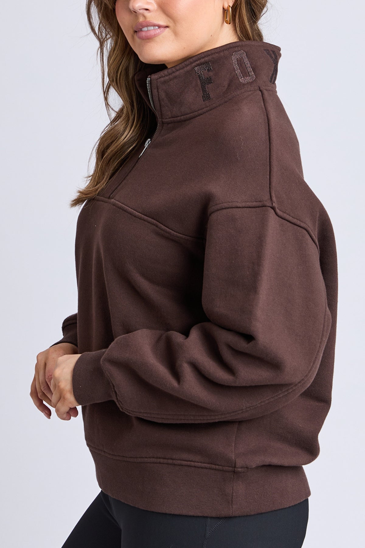 A woman models the Foxwood College Zip Crew sweatshirt in brown, showcasing "FOX" embroidery on the collar. She pairs it with black leggings and poses against a simple light background.