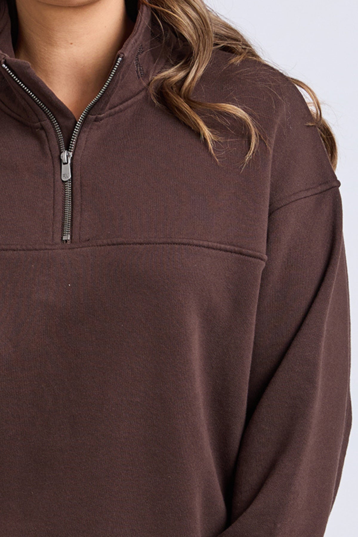 A person models the Foxwood College Zip Crew in brown, featuring a visible zipper, with wavy hair partially over their shoulder. The image is cropped from shoulders to midsection against a plain light background.