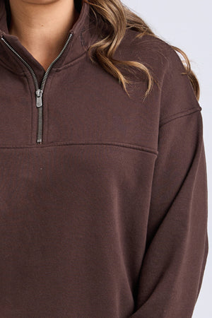 A person models the Foxwood College Zip Crew in brown, featuring a visible zipper, with wavy hair partially over their shoulder. The image is cropped from shoulders to midsection against a plain light background.