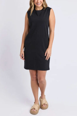 A woman smiles against a plain background in the knee-length, black Foxwood Reform Dress with a relaxed fit, styled with beige platform sandals and her wavy shoulder-length hair.