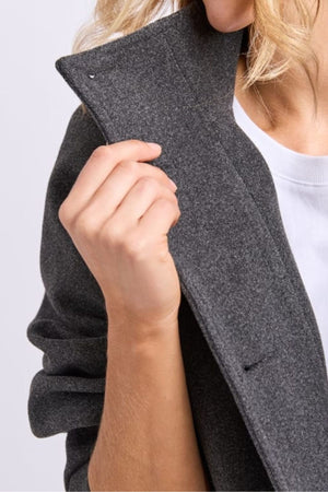 A woman with wavy blonde hair holds the collar of a dark gray Foxwood Fermont Coat, a chic transitional jacket. The cropped image highlights her upper torso, hand, and part of her face.