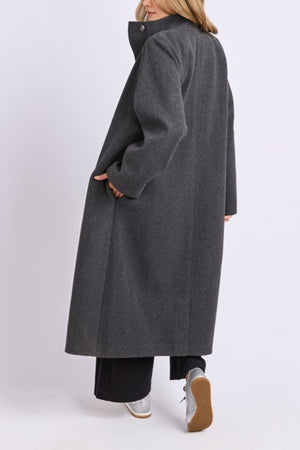 A woman models the long, dark gray Foxwood Fermont Coat with black pants and light sneakers, her back to the camera against a plain, light background.