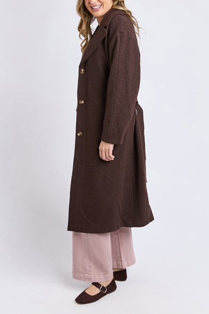 A person in a long, dark brown Foxwood Whistler Coat, light pink wide-leg pants, and brown shoes stands against a white background. The person's face is partially out of frame, and they are smiling.