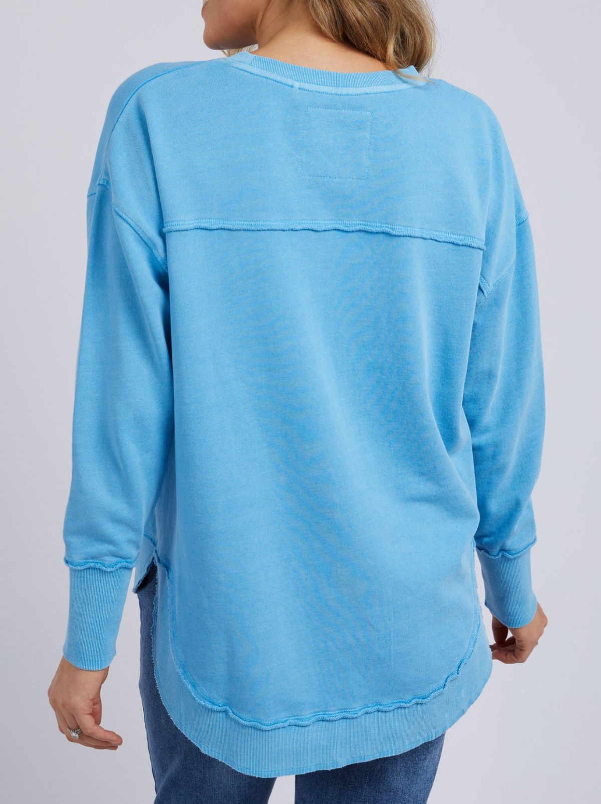 A person wears the Foxwood Simplified Crew, a loose-fitting, light blue long-sleeve jumper with visible seams, subtle Foxwood embroidery, and gently raw hems. The person has light brown hair.