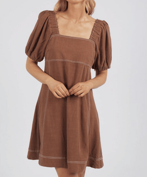 A person in the Foxwood Florence Dress, featuring a brown hue, puffed sleeves, and a square neckline, stands against a plain background. The dress's vertical seams highlight its relaxed fit and lightweight feel as the person gently clasps their hands in front.