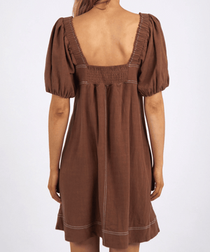 Against a plain backdrop, someone wears the Foxwood Florence Dress with its brown, puff-sleeves, square neckline, and back smocked details. The knee-length dress provides a lightweight comfort as their hair is tied back.