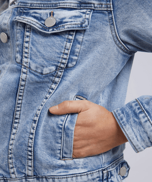 A person wears the Foxwood Georgia Denim Jacket, a smart-casual staple in light blue denim with silver buttons. The cotton-elastane blend has a subtle worn pattern, and their hand rests comfortably in the pocket against a neutral backdrop, emphasizing this versatile piece.
