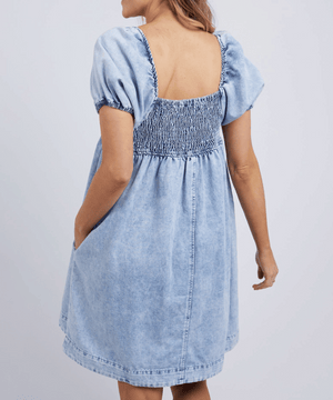 A woman is seen from behind wearing the Foxwood Jade Textured Dress, a light blue short-sleeve cotton dress with elastic smocking on the upper back and sleeves, a square neckline, vintage wash finish, and side pockets.