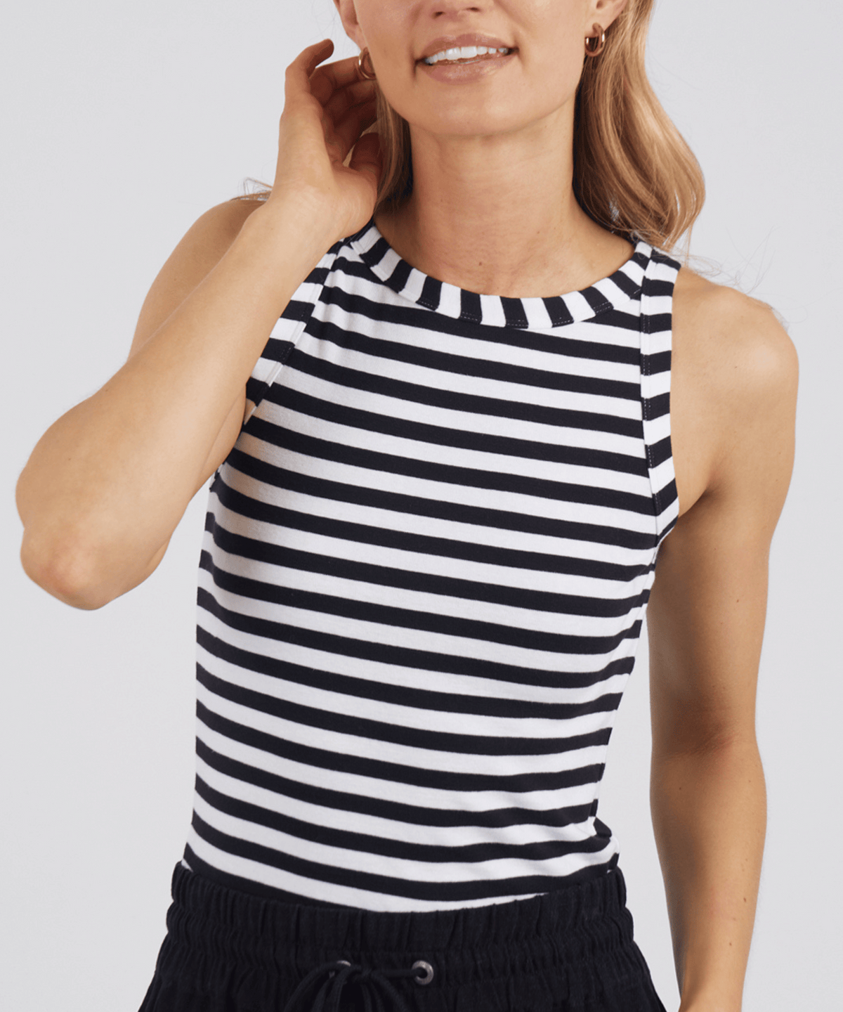 A woman wears the Foxwood Ruth Tank in black and white stripes with black pants, standing against a plain background, smiling gently with one hand at her neck.