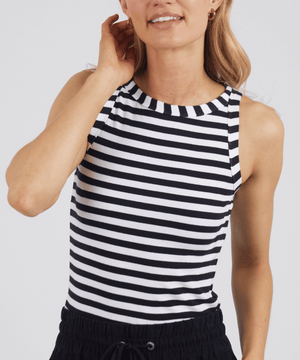 A woman wears the Foxwood Ruth Tank in black and white stripes with black pants, standing against a plain background, smiling gently with one hand at her neck.