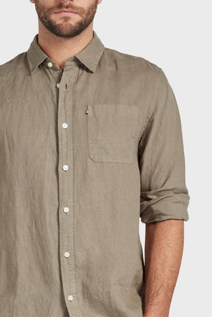 A man models the light brown Academy Brand Hampton Linen Shirt, featuring a front pocket, standing against a plain light background with only his upper body visible and his right arm partially out of frame.